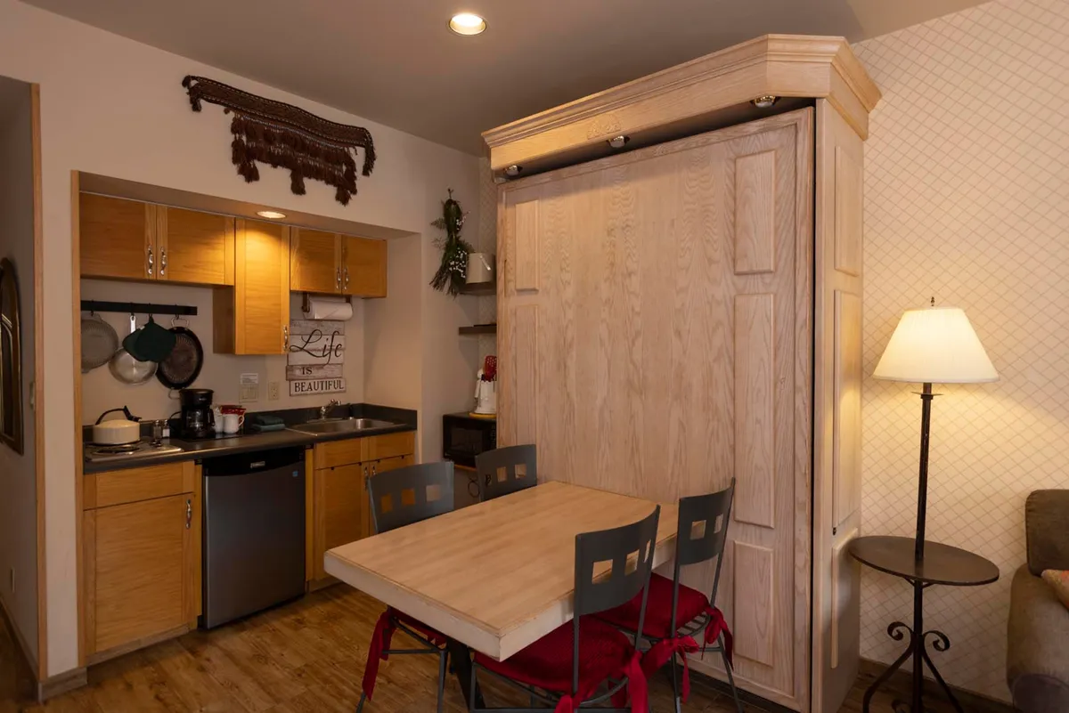 A compact kitchenette with modern appliances sits adjacent to a small dining table and a Murphy bed, maximizing space while remaining warm and inviting. The layout is practical yet cozy, perfect for preparing drinks or snacks while socializing. The ambiance encourages intimate dinners or casual gatherings, echoing the section's family-friendly, flexible vibe.