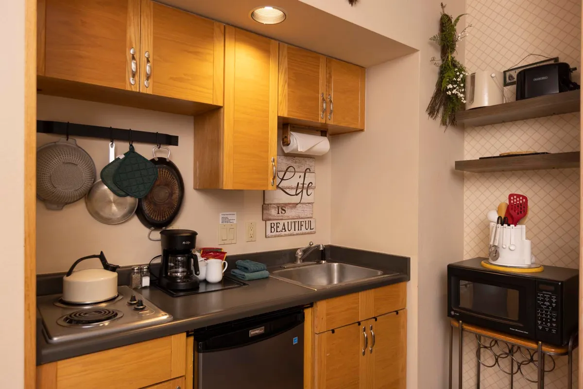 A warm, welcoming kitchen interior with a coffee maker on the countertop, decorative wall hanging, and space for casual dining. The scene suggests a lived-in atmosphere focused on daily conveniences and making small moments comfortable.