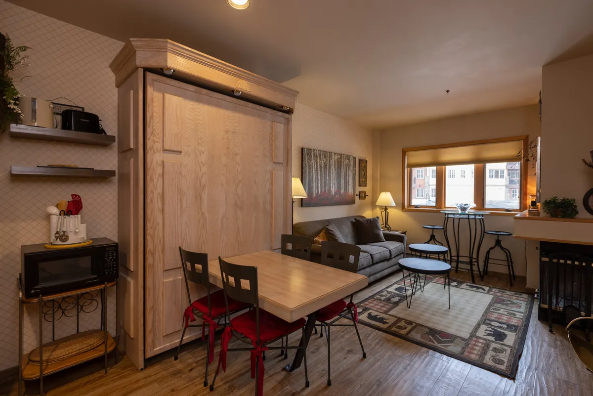 A comfortable, light-filled space with a folding wooden bed unit, a dining table with chairs, a cozy couch, and a large window letting in natural light. The arrangement suggests versatility—ideal for both daytime relaxation and evening gatherings. The table is perfect for games or meals, reinforcing the sense of togetherness central to the section's narrative.