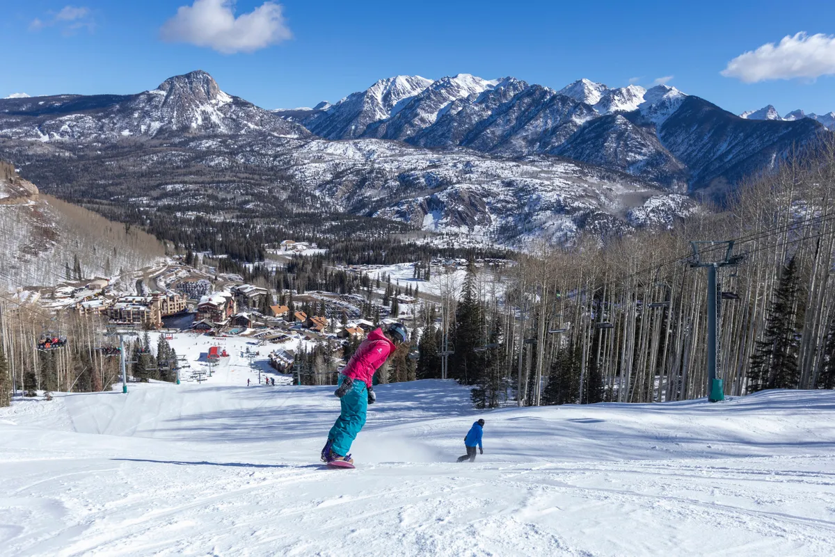 A vibrant winter scene with a skier in bright clothing and a child skiing, set against snow-covered mountains and resort buildings. The joyful activity and family presence underscore the fun and dynamic atmosphere of the resort.