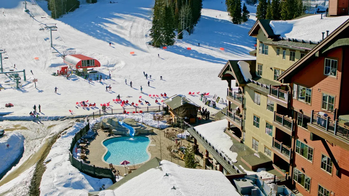 This exterior shot captures an outdoor pool with views of the ski slopes, ski lift, and resort buildings. The area is bustling with guests enjoying the pool and socializing, while the surrounding mountain setting adds a lively, communal vibe.