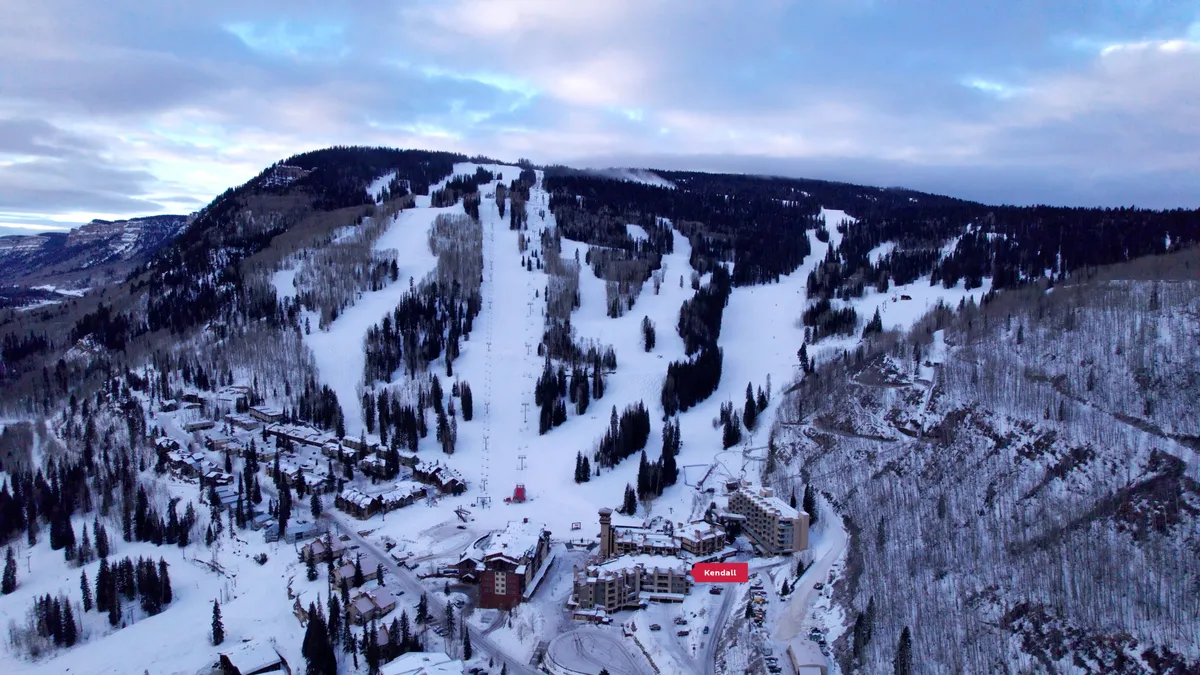 This exterior image captures the vibrant resort atmosphere at Kendall 233, with snow-covered mountains in the background, ski slopes bustling with activity, and resort buildings nestled at the base. Guests can be seen enjoying skiing and snowboarding, while others relax near the cozy lodge, evoking a sense of adventure and togetherness.