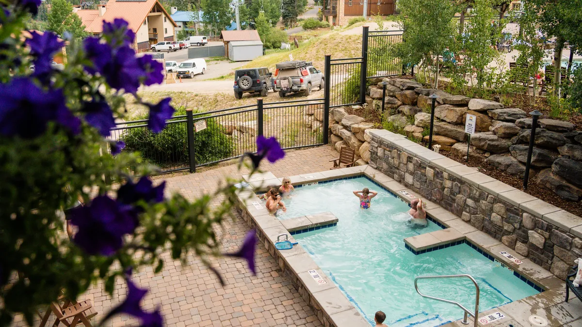 A vibrant outdoor swimming pool is shown, bordered by flowers and set against a lush landscape. Sunlight glimmers off the water, and guests are seen swimming, sunbathing, and socializing in a communal, joyful setting.
