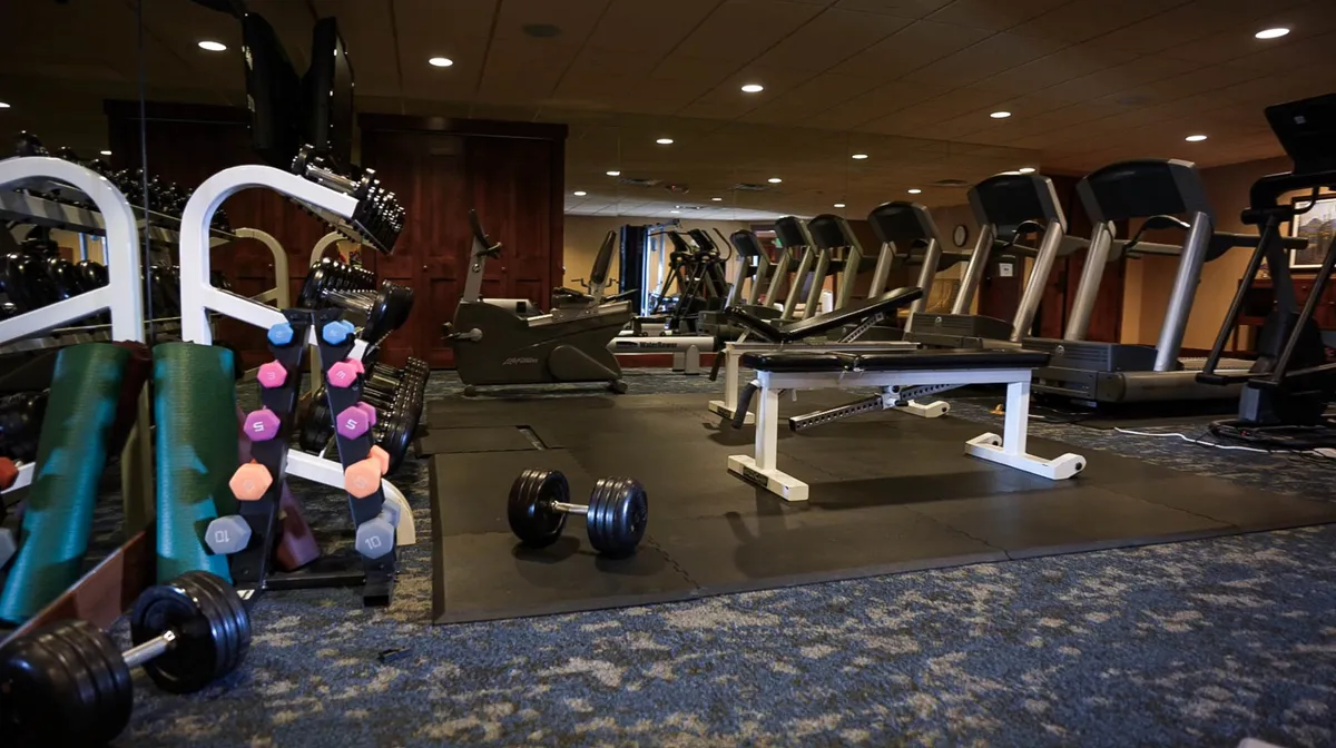 This image captures a modern fitness center with well-maintained dumbbells, treadmills, a weight bench, and various exercise machines. The space is brightly lit, energetic, and arranged to support both individual workouts and group fitness sessions, reflecting a welcoming, community-oriented environment.