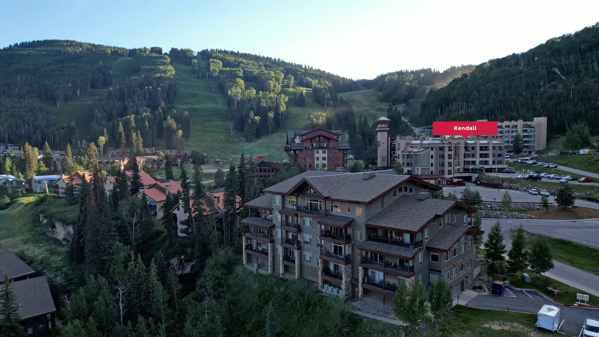 An exterior view of the resort buildings set against a lush mountain landscape, with visible parking areas and immediate access to the Village Plaza. The image captures the seamless blend of private sanctuary and vibrant resort surroundings.