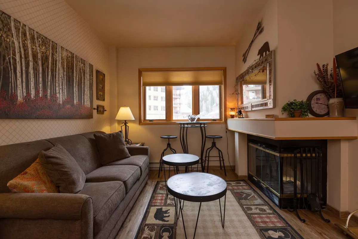 The image showcases a cozy living area centered around a glowing wood-burning fireplace, with a comfortable sofa, modern coffee tables, tasteful artwork, and a large window framing views of the snowy Colorado landscape. The space feels warm and inviting, perfect for relaxing or sharing stories after a day on the mountain.