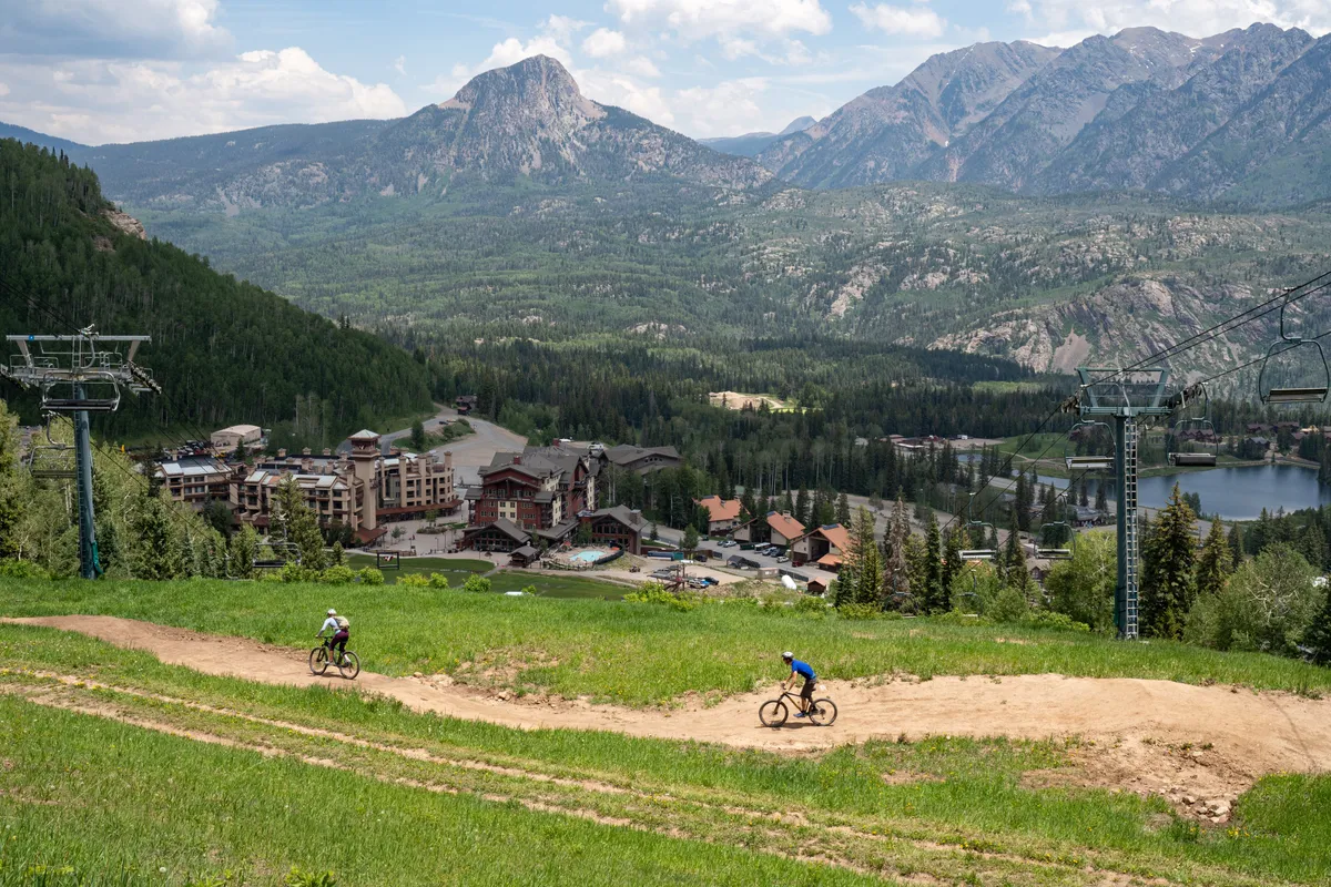 This exterior shot captures the mountain range, bicyclists on a trail, resort buildings, and a ski lift. The scene reflects year-round adventure, showcasing mountain biking, hiking, and the proximity of the resort to outdoor experiences.