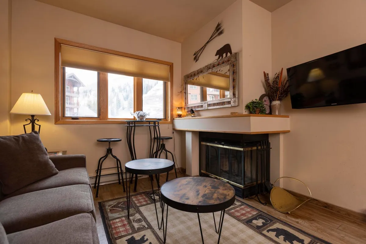 A welcoming living area featuring a plush sofa arranged around a wood-burning fireplace. Bar stools are visible at the counter, suggesting a seamless connection between the living space and the adjacent kitchenette. Decorative touches like a mirror and cozy decor enhance the feeling of warmth and invitation. Through the window, snowy scenery hints at the mountain setting. Guests are seen relaxing, socializing, and enjoying hot drinks together.