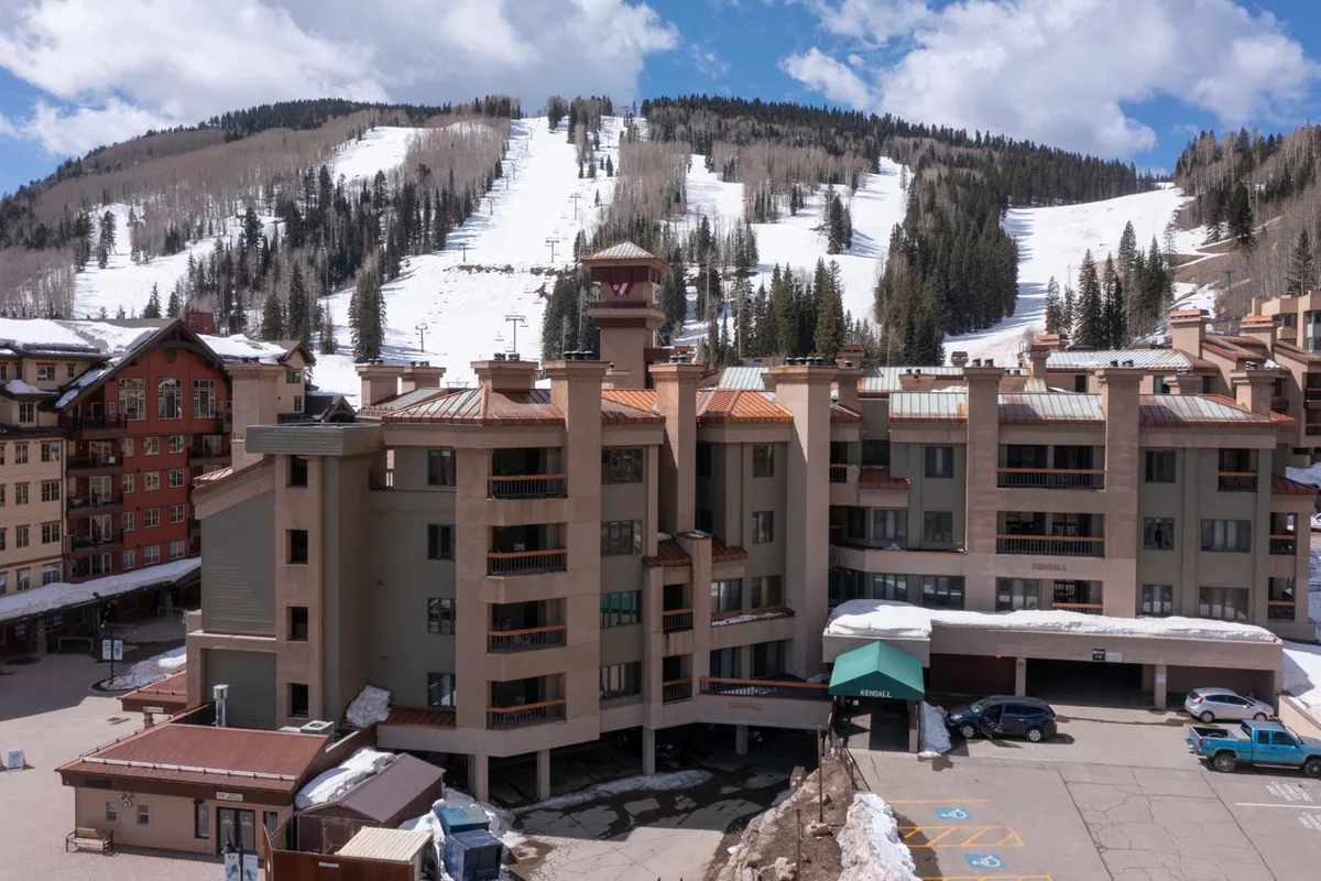 This image features the exterior of the building, with ski slopes and surrounding trees visible in the background. The scene includes hints of activity around the parking area, conveying proximity to adventure and easy access to the slopes. The overall mood is adventurous yet cozy, with a clear sense of being at the heart of the mountain environment.