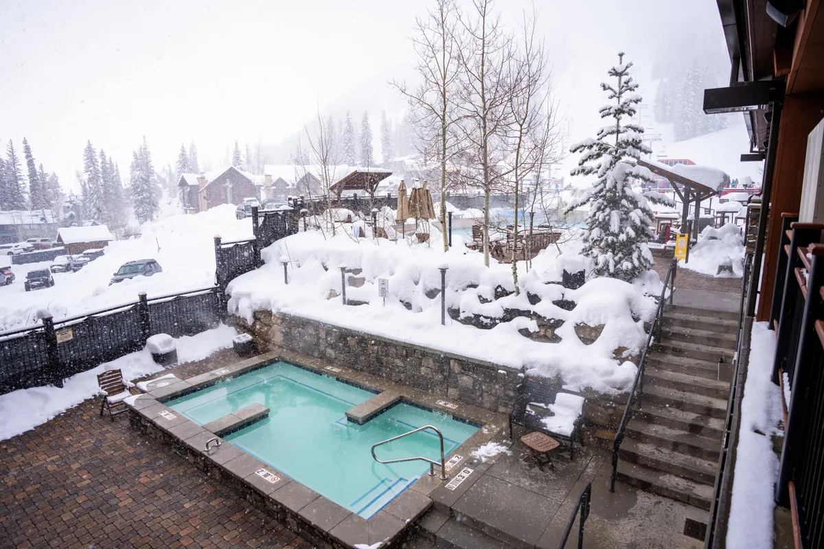 This image features a hot tub set amidst a snow-covered landscape, with steps leading up to it and surrounded by tall trees. People are relaxing in the bubbling water, enveloped by winter scenery, creating a tranquil and rejuvenating mood.